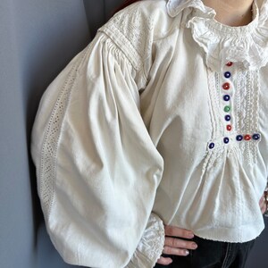 Op de afbeelding: Een wit linnen blouse met gepofte mouwen en een gerimpelde kraag. De blouse heeft ingewikkelde kantdetails en kleurrijke knopen aan de voorkant.