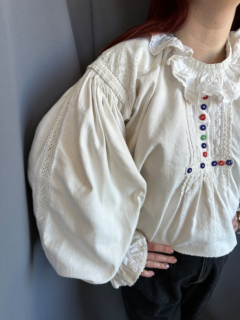 Op de afbeelding: Een wit linnen blouse met gepofte mouwen en een gerimpelde kraag. De blouse heeft ingewikkelde kantdetails en kleurrijke knopen aan de voorkant.