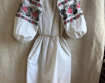 May include: White linen dress with long, puffed sleeves. The sleeves and hem are adorned with intricate embroidery in red and black floral patterns. A simple tie belt cinches the waist. The dress hangs on a hanger against a neutral background.