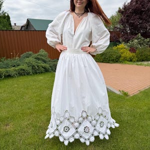 May include: A white dress with long sleeves and a deep V-neck. The dress features embroidered details on the sleeves and a decorative, openwork hem. The woman is standing on green grass.