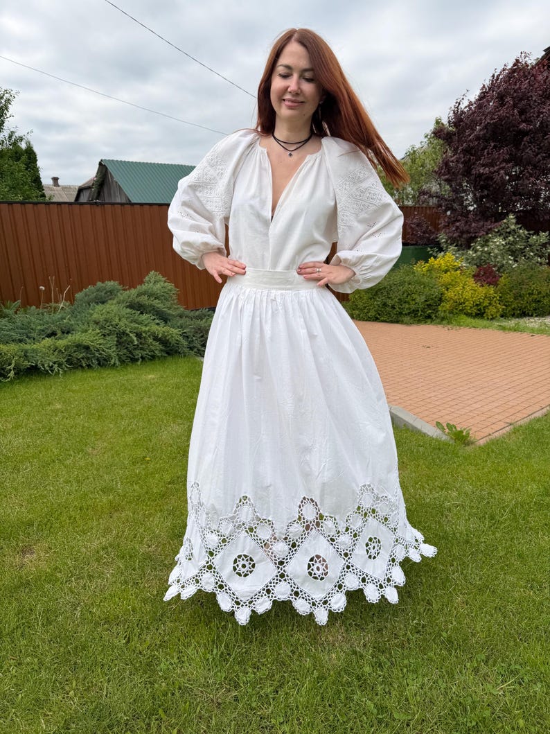 May include: A white dress with long sleeves and a deep V-neck. The dress features embroidered details on the sleeves and a decorative, openwork hem. The woman is standing on green grass.
