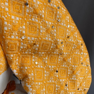 May include: A white linen blouse with a yellow embroidered design and silver sequins. The embroidery is a geometric pattern of squares and diamonds.