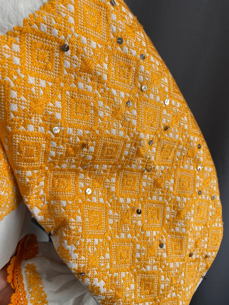 May include: A white linen blouse with a yellow embroidered design and silver sequins. The embroidery is a geometric pattern of squares and diamonds.