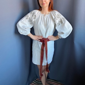 May include: A white linen tunic featuring long, voluminous sleeves with grey floral embroidery on the shoulders and a round neckline. A long, woven belt with vertical stripes in red, yellow, and dark blue is tied around the waist with fringed ends.