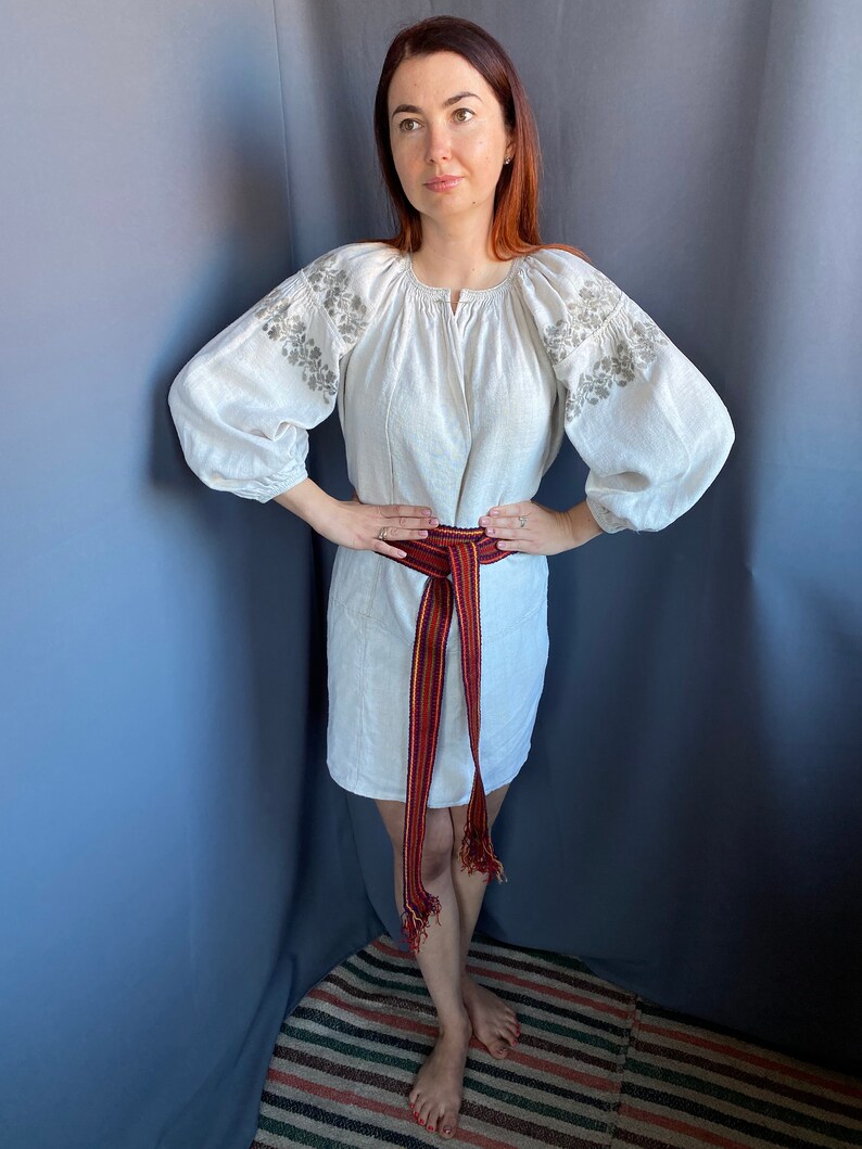 May include: A white linen tunic featuring long, voluminous sleeves with grey floral embroidery on the shoulders and a round neckline. A long, woven belt with vertical stripes in red, yellow, and dark blue is tied around the waist with fringed ends.