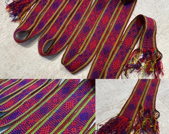 May include: A close-up of a red, blue, and yellow woven fabric with fringe. The fabric is made of a variety of different threads and has a geometric pattern.