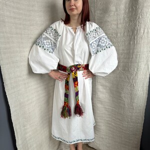May include: A white linen dress with embroidered sleeves and a colorful woven belt. The dress has a loose fit and a round neckline. The belt is tied at the waist and has long tassels.