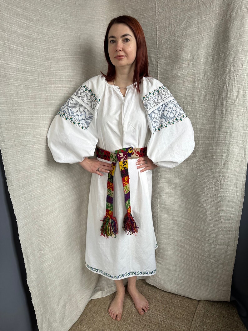 May include: A white linen dress with embroidered sleeves and a colorful woven belt. The dress has a loose fit and a round neckline. The belt is tied at the waist and has long tassels.