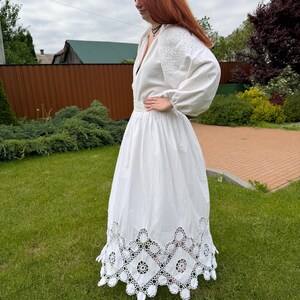 May include: A white dress with long sleeves and intricate embroidery on the bodice and sleeves. The dress features a deep V-neck and a full skirt with a decorative, openwork hem. The dress is made of a lightweight fabric.