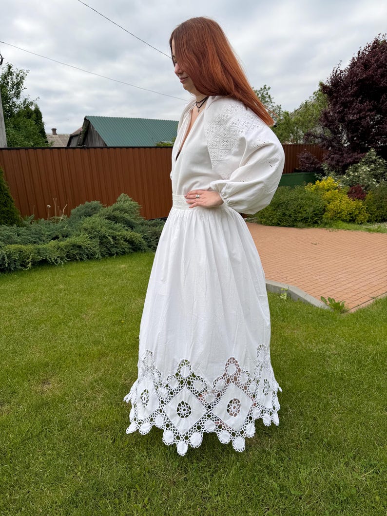 May include: A white dress with long sleeves and intricate embroidery on the bodice and sleeves. The dress features a deep V-neck and a full skirt with a decorative, openwork hem. The dress is made of a lightweight fabric.