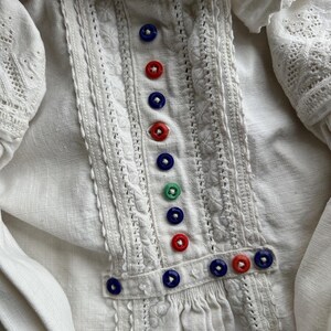 Op de afbeelding: Wit linnen blouse met delicate kantdetails en een rij kleurrijke knopen aan de voorkant.