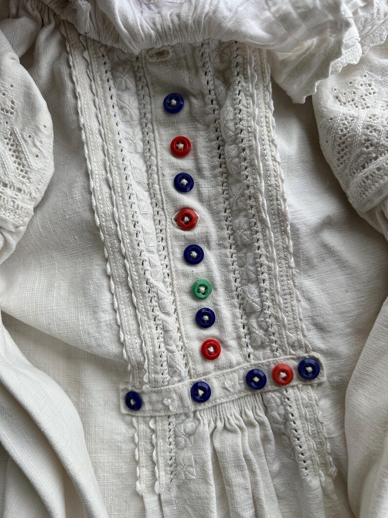 Op de afbeelding: Wit linnen blouse met delicate kantdetails en een rij kleurrijke knopen aan de voorkant.