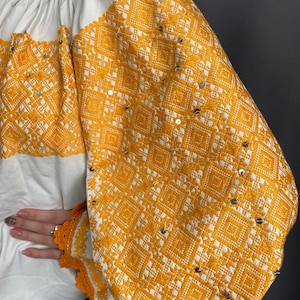 May include: White blouse with long sleeves and a yellow embroidered pattern. The embroidery features a geometric design with small silver sequins.