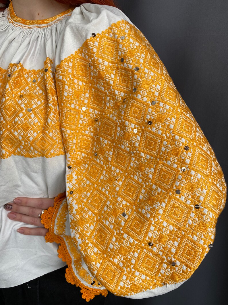 May include: White blouse with long sleeves and a yellow embroidered pattern. The embroidery features a geometric design with small silver sequins.