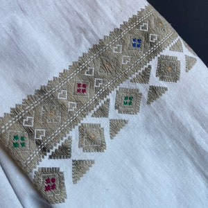May include: White linen fabric with a gray embroidered design. The embroidery features geometric shapes and small squares in red, green, and blue.
