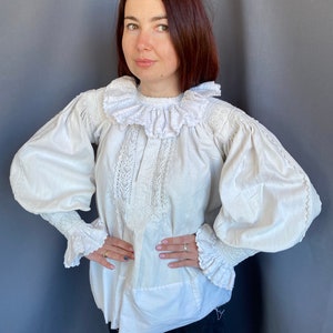 K&ouml;nnte beinhalten: Eine wei&szlig;e Bluse mit einem gro&szlig;en, gekr&auml;uselten Kragen und langen, bauschigen &Auml;rmeln. Die &Auml;rmel haben an den Manschetten Spitzenbesatz. Die Bluse ist aus einem leichten Stoff gefertigt und hat eine lockere Passform.