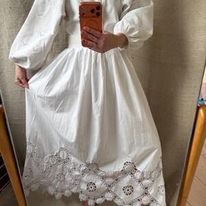 May include: White cotton dress with long, puffy sleeves and intricate eyelet embroidery on the sleeves and skirt hem. The dress has a V-neckline and a full skirt, creating a flowing silhouette. The hem features a detailed, openwork lace design.