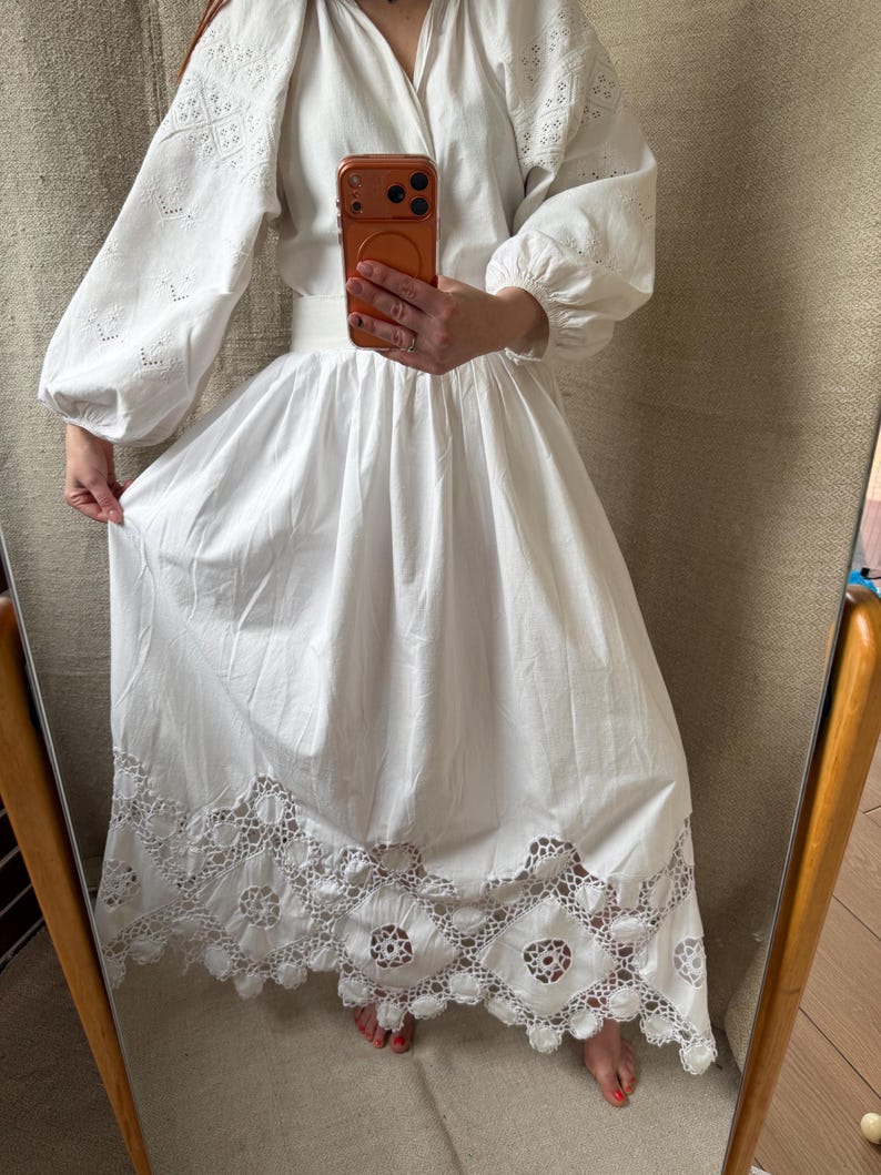 May include: White cotton dress with long, puffy sleeves and intricate eyelet embroidery on the sleeves and skirt hem. The dress has a V-neckline and a full skirt, creating a flowing silhouette. The hem features a detailed, openwork lace design.