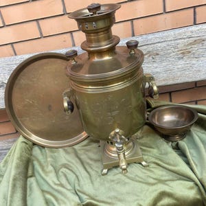 May include: A brass samovar featuring a central urn with a spigot, two side handles with dark wooden accents, and a top lid. The set includes a matching circular brass tray and a small brass bowl, all displaying a polished metallic finish.