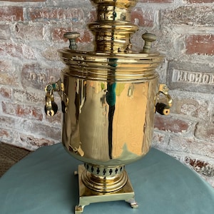 May include: A large, antique brass samovar with a round body, two handles, and a square base. The samovar has a tall, cylindrical spout with a lid on top. The samovar is sitting on a green tablecloth.