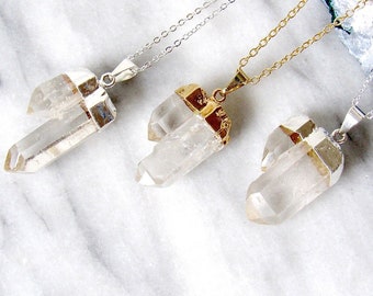 May include: Three clear quartz crystal necklaces with silver and gold-colored metal accents. Each crystal is suspended from a delicate chain. The necklaces are displayed on a white and gray marble surface.