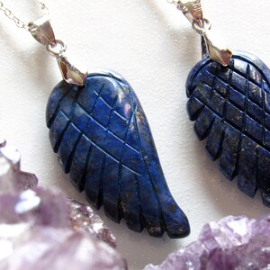 May include: Two lapis lazuli wing pendants with silver-colored chains. The wings are intricately carved with feather details. The pendants are a deep blue color, and the background is white with purple crystals.