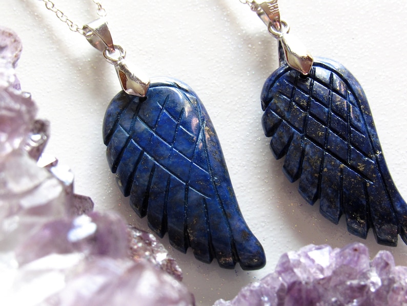 May include: Two lapis lazuli wing pendants with silver-colored chains. The wings are intricately carved with feather details. The pendants are a deep blue color, and the background is white with purple crystals.