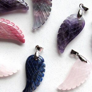 May include: Assortment of gemstone wing pendants in various colors, including purple, pink, and blue. Each pendant features intricate carved feather details and a silver-colored bail for attaching to a chain. The pendants are displayed on a white speckled surface.