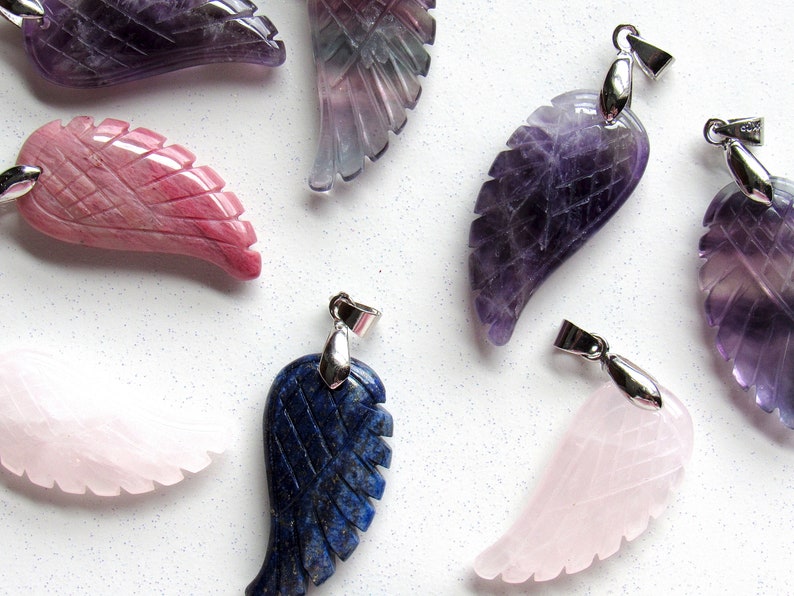 May include: Assortment of gemstone wing pendants in various colors, including purple, pink, and blue. Each pendant features intricate carved feather details and a silver-colored bail for attaching to a chain. The pendants are displayed on a white speckled surface.