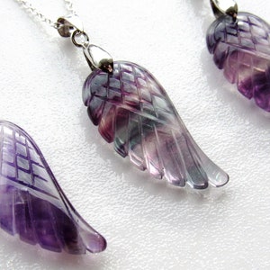 May include: Close-up of three gemstone wing pendants on silver chains. The wings are carved from purple, green, and clear fluorite. The pendants are displayed on a white surface. Jewelry.