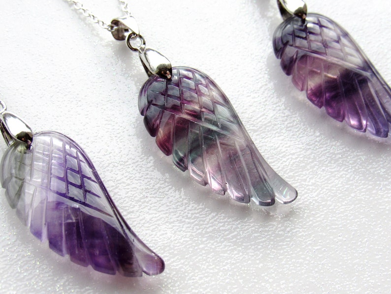 May include: Close-up of three gemstone wing pendants on silver chains. The wings are carved from purple, green, and clear fluorite. The pendants are displayed on a white surface. Jewelry.
