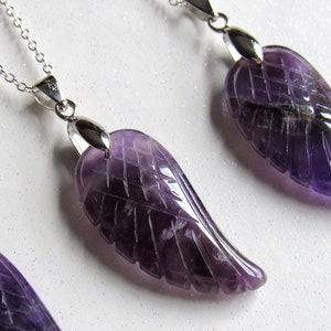 May include: Amethyst wing pendant necklaces with silver chains. The purple gemstone wings are carved with feather details. The necklaces are displayed on a white, glittery surface.