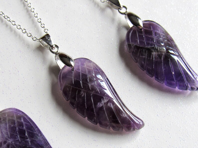 May include: Amethyst wing pendant necklaces with silver chains. The purple gemstone wings are carved with feather details. The necklaces are displayed on a white, glittery surface.