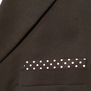 May include: Close-up of a black blazer with a white polka dot pocket square. The pocket square is rectangular and features evenly spaced white dots on a black background. The blazer's pocket is visible.