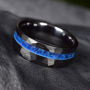 Puede incluir: Un anillo de carburo de tungsteno negro con una incrustaci&oacute;n de purpurina azul. El anillo tiene un acabado martillado.