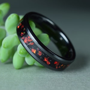 Könnte beinhalten: Ein schwarzer Ring mit einem Band aus roten und orangen Edelsteinen. Der Ring ist vor dem Hintergrund grüner Sukkulenten und einer grauen Oberfläche platziert. Schmuck.