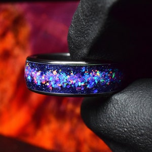 May include: A black ring with a blue band filled with iridescent glitter. The glitter is a mix of pink, blue, green, and yellow.