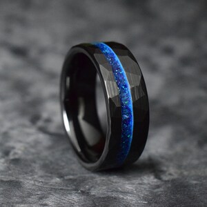 Puede incluir: Un anillo de carburo de tungsteno negro con una incrustaci&oacute;n azul y negra. El anillo tiene un acabado martillado.