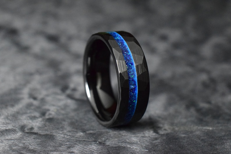 Puede incluir: Un anillo de carburo de tungsteno negro con una incrustaci&oacute;n azul y negra. El anillo tiene un acabado martillado.