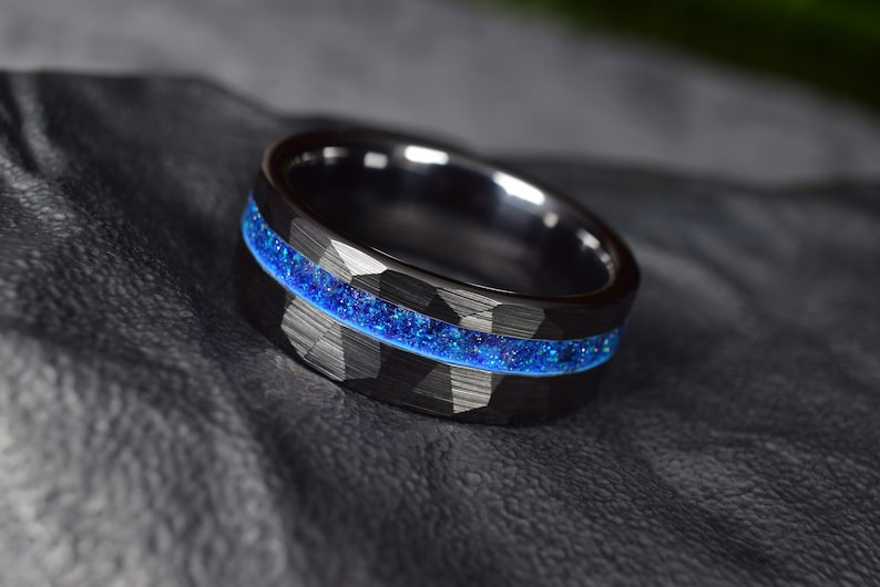 Puede incluir: Un anillo de carburo de tungsteno negro con una incrustaci&oacute;n de purpurina azul. El anillo tiene un acabado martillado.
