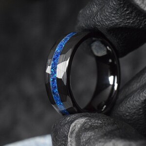 Puede incluir: Un anillo de carburo de tungsteno negro con una incrustaci&oacute;n de purpurina azul. El anillo tiene un borde biselado y un acabado pulido.