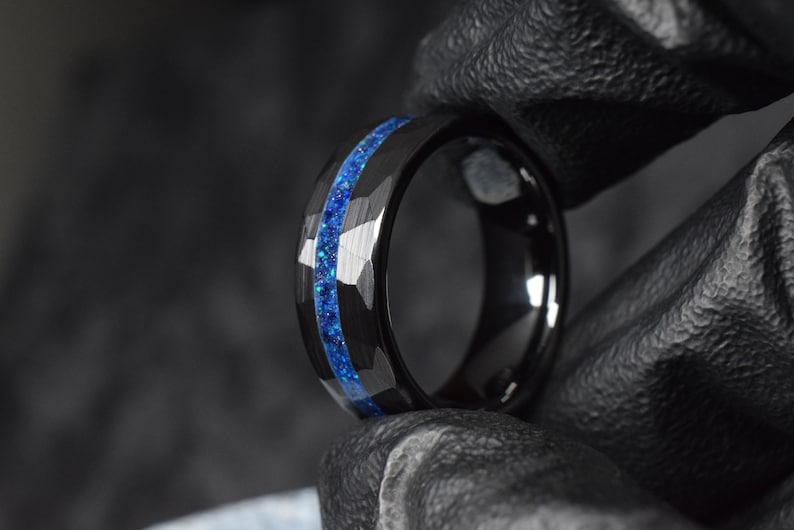 Puede incluir: Un anillo de carburo de tungsteno negro con una incrustaci&oacute;n de purpurina azul. El anillo tiene un borde biselado y un acabado pulido.