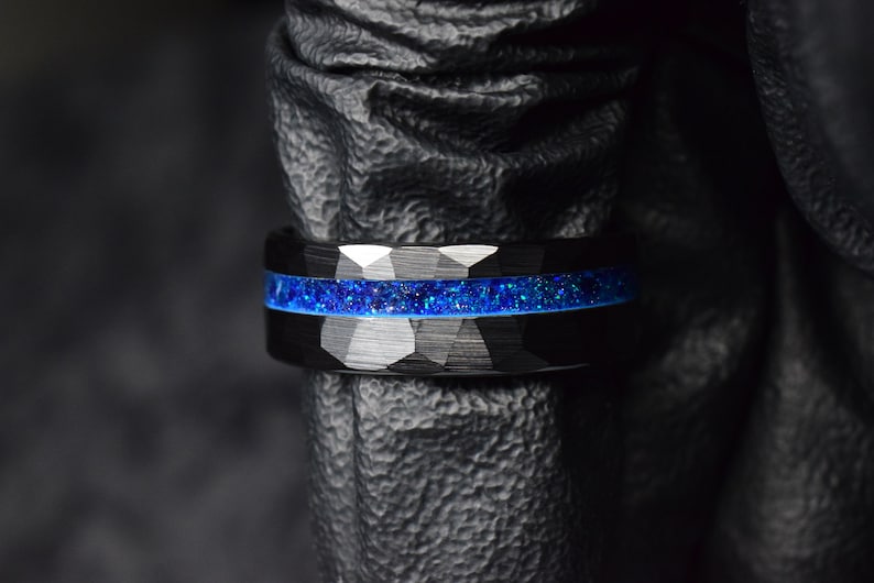 Puede incluir: Un anillo de carburo de tungsteno negro con una incrustaci&oacute;n de purpurina azul. El anillo tiene un acabado cepillado y un borde biselado.