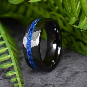 Puede incluir: Un anillo de carburo de tungsteno negro con una incrustaci&oacute;n de purpurina azul. El anillo tiene un borde biselado y un acabado pulido.