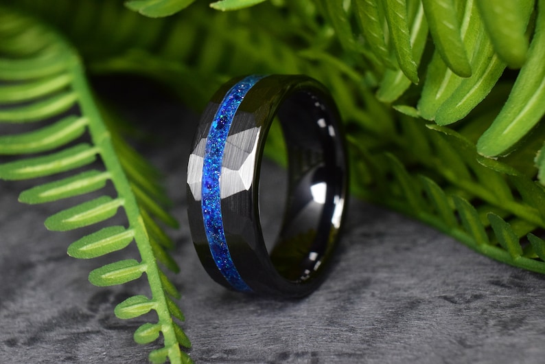 Puede incluir: Un anillo de carburo de tungsteno negro con una incrustaci&oacute;n de purpurina azul. El anillo tiene un borde biselado y un acabado pulido.