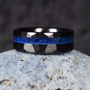 Puede incluir: Un anillo de carburo de tungsteno negro con una incrustaci&oacute;n de purpurina azul. El anillo tiene un acabado cepillado y un borde biselado.