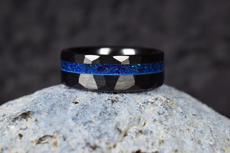 Puede incluir: Un anillo de carburo de tungsteno negro con una incrustaci&oacute;n de purpurina azul. El anillo tiene un acabado cepillado y un borde biselado.