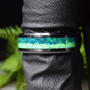 May include: A black tungsten carbide ring with a green and blue opal inlay. The ring is on a dark gray surface.