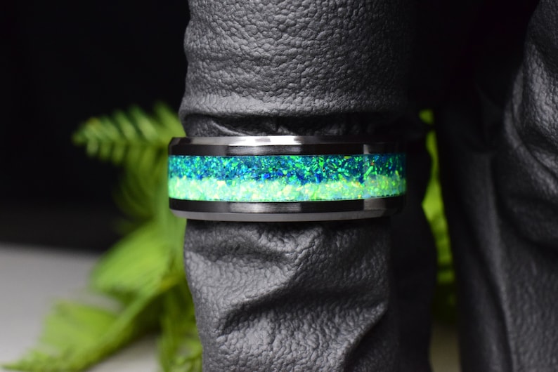 May include: A black tungsten carbide ring with a green and blue opal inlay. The ring is on a dark gray surface.