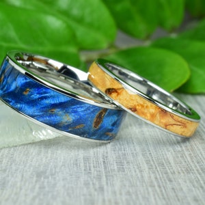 Könnte beinhalten: Zwei Eheringe mit einem silbernen Metallband und einer Holzeinlage. Ein Ring hat eine blaue Holzeinlage und der andere eine hellbraune Holzeinlage.